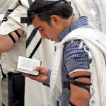 Jewish men wearing tallitot (prayer shawls) pray with tefillin (phylacteries) and siddurim (Jewish prayer books).