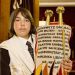 A Jewish teen holds a Torah scroll covered by an ornately decorated Torah mantle that commemorates those who perished in Nazi death camps during the Holocaust.