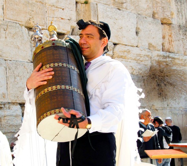 A Jewish man wearing tefillin (phylacteries) holds a Torah scroll protected by a study case called a tik.