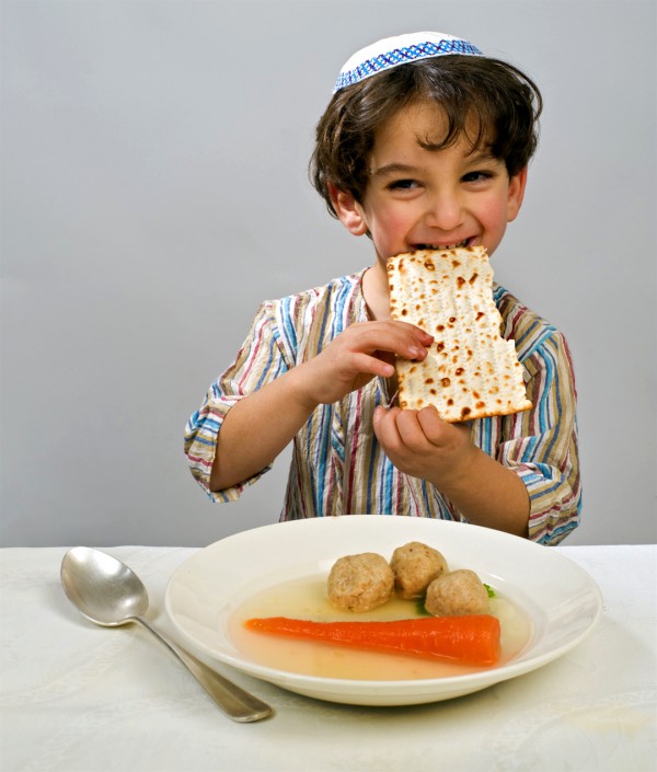 A Jewish boy eats a piece of matzah. Before him is a bowl of matzah ball soup.