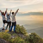 three people stand on mountain top raising hands in praise.