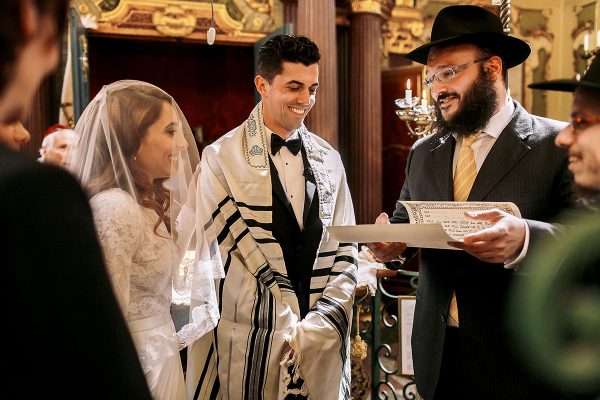 Jewish bride stands next to Jewish groom wearing a tallit (prayer shawl).  